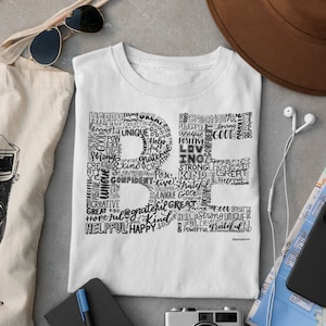 Puede incluir: Una camiseta blanca con un dise&ntilde;o gr&aacute;fico negro en forma de la letra "R" compuesta por palabras como "feliz", "fuerte", "genial", "amor", "amable", "&uacute;nico", "servicial" y "agradecido".