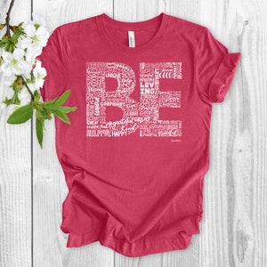 Puede incluir: Una camiseta roja con un dise&ntilde;o gr&aacute;fico blanco que deletrea "BE" usando palabras positivas como "feliz", "fuerte", "amable" y "agradecido".