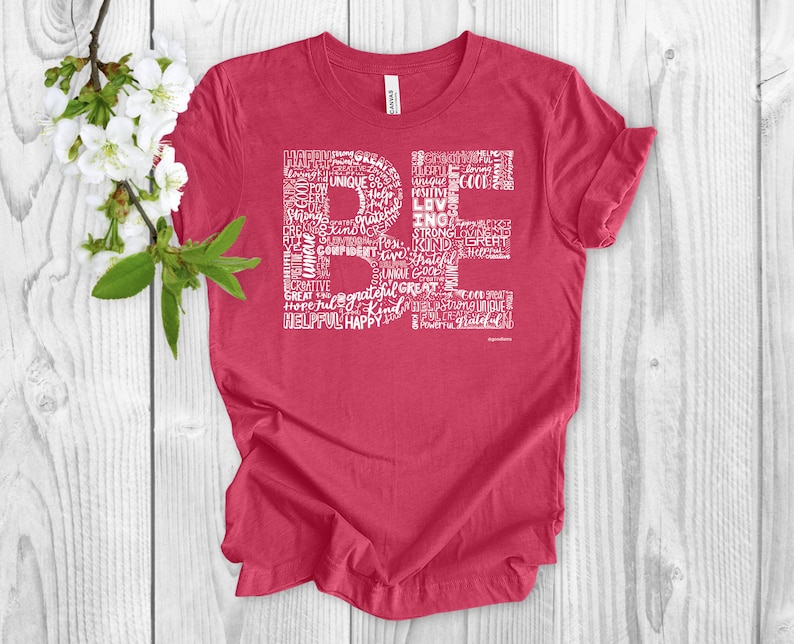 Puede incluir: Una camiseta roja con un dise&ntilde;o gr&aacute;fico blanco que deletrea "BE" usando palabras positivas como "feliz", "fuerte", "amable" y "agradecido".