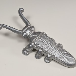 May include: A silver-colored metal beetle-shaped object with two long antennae. The back of the beetle features embossed text that reads "CENTENNIAL" and "LINCOLN 1867". The surface has a textured, cast metal appearance with visible legs on the sides.