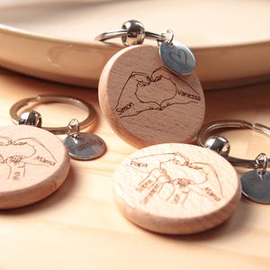 K&ouml;nnte beinhalten: Drei Holzschl&uuml;sselanh&auml;nger mit gravierten Handgesten, die ein Herz bilden. Jeder Schl&uuml;sselanh&auml;nger tr&auml;gt einen anderen Namen: "Simon & Vanessa", "Familie" und "Papa, Sophia, Daniel & Mama".