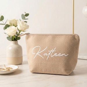 Puede incluir: Un neceser beige con el nombre "Katteen" en escritura blanca. La bolsa tiene cierre de cremallera y est&aacute; hecha de un material suave. Un peque&ntilde;o jarr&oacute;n con rosas blancas y un plato con joyas de oro est&aacute;n en el fondo.