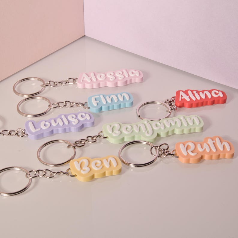K&ouml;nnte beinhalten: Eine Auswahl an bunten Schl&uuml;sselanh&auml;ngern mit Namen wie "Alessia" und "Finn" in verspielter Schrift. Jeder Schl&uuml;sselanh&auml;nger hat eine andere Pastellfarbe, darunter Rosa, Blau und Gr&uuml;n, mit einer silbernen Kette und einem Schl&uuml;sselring.