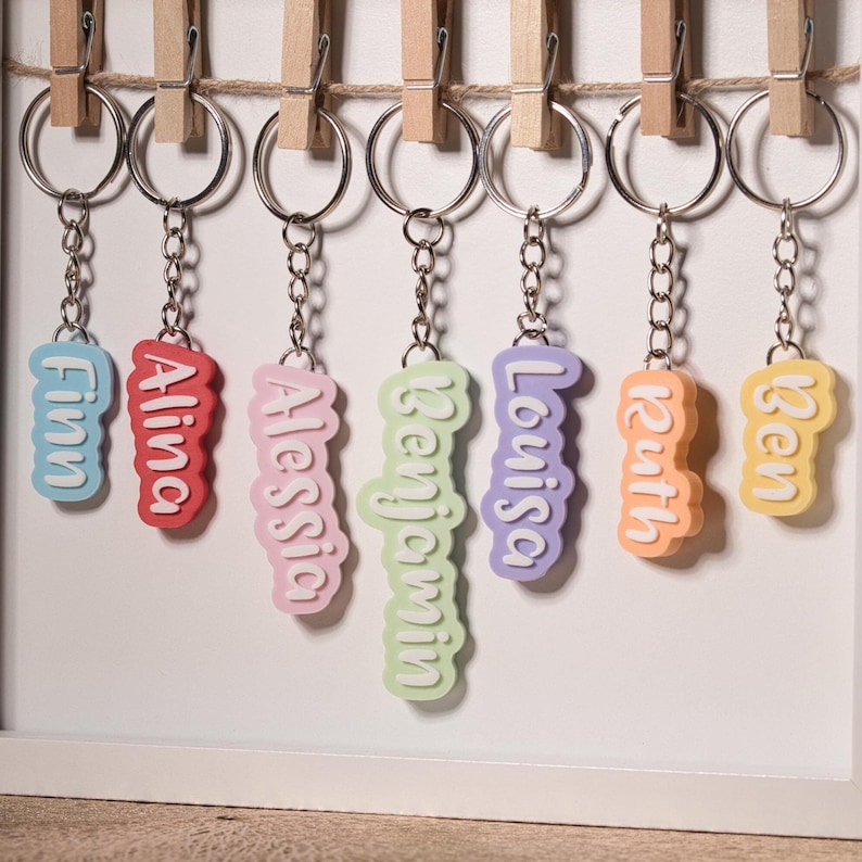 K&ouml;nnte beinhalten: Eine Sammlung farbenfroher Namensanh&auml;nger, jeder mit einem anderen Namen in einer verspielten Schriftart. Die Schl&uuml;sselanh&auml;nger sind in verschiedenen Farben erh&auml;ltlich, darunter Blau, Rot, Rosa, Gr&uuml;n, Lila, Orange und Gelb. Sie h&auml;ngen an silbernen Schl&uuml;sselringen.