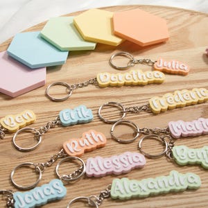 K&ouml;nnte beinhalten: Eine Sammlung farbenfroher Namens-Schl&uuml;sselanh&auml;nger in Pastellt&ouml;nen, darunter Rosa, Blau, Gelb und Orange. Jeder Schl&uuml;sselanh&auml;nger zeigt einen anderen Namen in einer verspielten Schriftart, befestigt an einer silbernen Kette und einem Schl&uuml;sselring. Sechseckige Pastelldekorationen sind ebenfalls vorhanden.