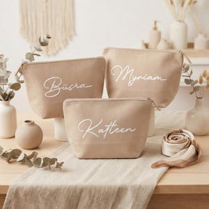 Puede incluir: Tres neceseres de lona beige con nombres en cursiva blanca: B&uuml;sra, Myriam y Katleen. Cada bolsa tiene un cierre de cremallera. Las bolsas se exhiben sobre una superficie de color claro con elementos decorativos en el fondo.