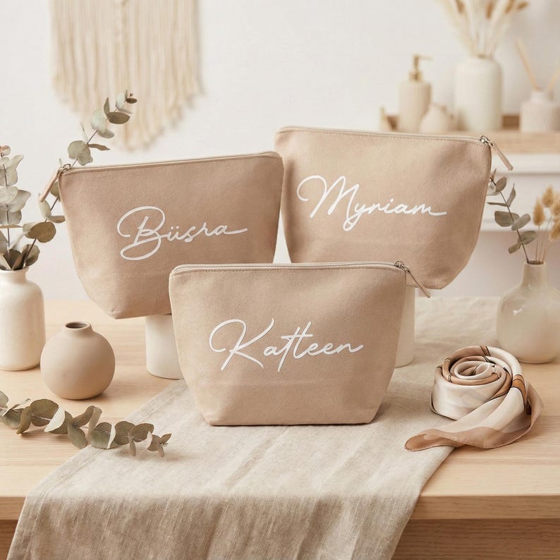 Puede incluir: Tres neceseres de lona beige con nombres en cursiva blanca: B&uuml;sra, Myriam y Katleen. Cada bolsa tiene un cierre de cremallera. Las bolsas se exhiben sobre una superficie de color claro con elementos decorativos en el fondo.