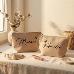 Puede incluir: Dos neceseres de lona beige con texto cursivo negro que dice "Marina" y "Amelie", cada uno con un peque&ntilde;o coraz&oacute;n negro. Un espejo con marco dorado, un tubo de brillo labial y un plato peque&ntilde;o con anillos est&aacute;n sobre la mesa.