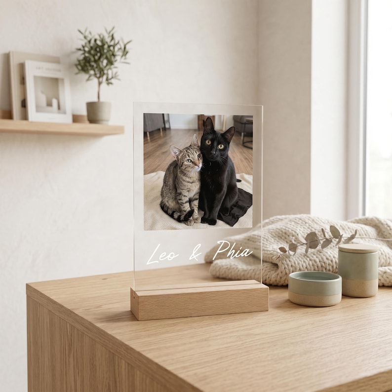 Peut inclure: Un cadre photo en acrylique transparent avec une base en bois affiche une photo de deux chats, l'un tigr&eacute; et l'autre noir. Les noms "Leo & Phia" sont &eacute;crits en script en bas. Le cadre est pos&eacute; sur une table en bois.