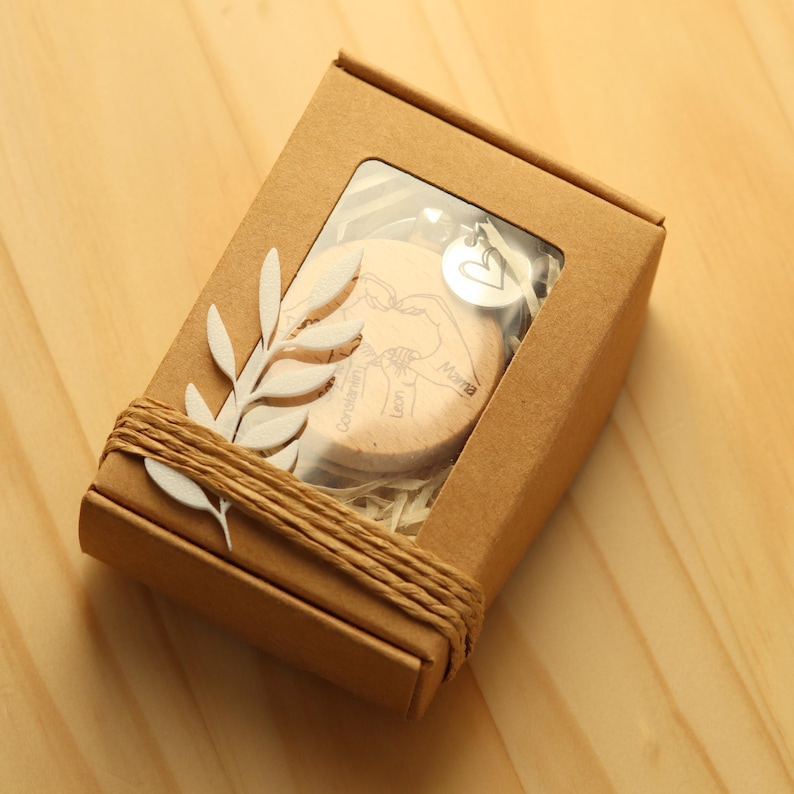 K&ouml;nnte beinhalten: Eine kleine Holzkiste mit einem durchsichtigen Fenster, die ein herzf&ouml;rmiges Holz-Ornament mit den eingravierten Worten "Gl&uuml;ckwunsch" und "Liebe" enth&auml;lt. Das Ornament wird von einem silbernen Herz-Anh&auml;nger begleitet.