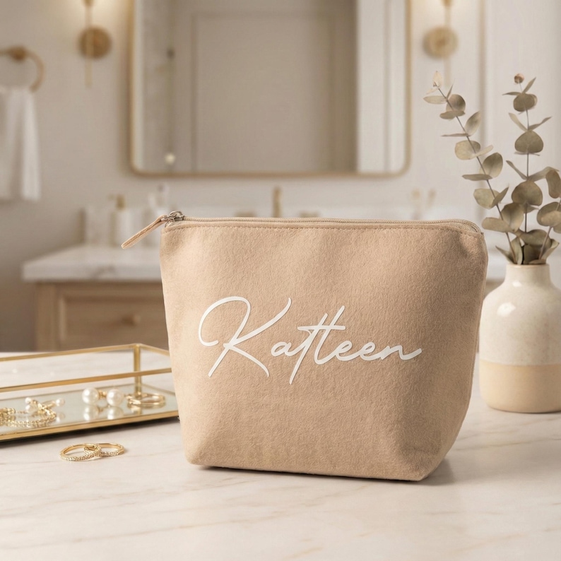 Puede incluir: Un neceser beige con el nombre "Katteen" en escritura blanca. La bolsa tiene un cierre de cremallera y est&aacute; sobre una superficie de m&aacute;rmol. El fondo incluye un tocador de ba&ntilde;o, espejo y objetos decorativos.