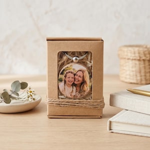 K&ouml;nnte beinhalten: Ein runder Holzschmuck mit einem Foto einer Frau und eines Kindes wird in einer Pappschachtel pr&auml;sentiert. Die Schachtel ist mit Bindfaden gebunden und hat ein Fenster, das den Schmuck zeigt. Der Schmuck hat eine silberne Metallschlaufe.