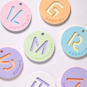 May include: Six pastel-coloured round name tags with a unique letter design and a name printed below. The colours are light pink, orange, light blue, light green, purple, and white.
