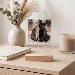 Peut inclure: Cadre photo en acrylique transparent avec une base en bois, affichant une photo de deux chats. L'un est noir, l'autre tigr&eacute;. Le texte "Leo & Phia" est grav&eacute; au bas du cadre. Le cadre est sur une surface en bois.