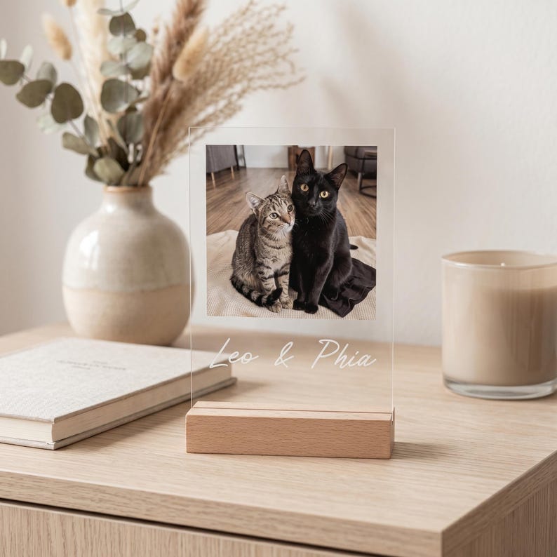 Peut inclure: Cadre photo en acrylique transparent avec une base en bois, affichant une photo de deux chats. L'un est noir, l'autre tigr&eacute;. Le texte "Leo & Phia" est grav&eacute; au bas du cadre. Le cadre est sur une surface en bois.