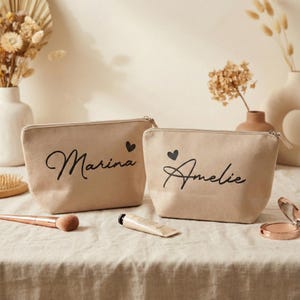 Puede incluir: Dos neceseres de lona beige con texto negro en cursiva. Una bolsa dice "Marina" y la otra "Amelie", cada una con un peque&ntilde;o coraz&oacute;n negro. Las bolsas est&aacute;n sobre una superficie de tonos neutros con brochas de maquillaje y un tubo de crema.