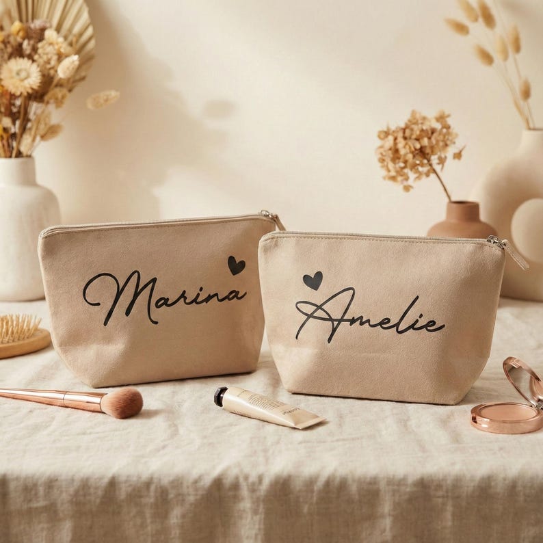 Puede incluir: Dos neceseres de lona beige con texto negro en cursiva. Una bolsa dice "Marina" y la otra "Amelie", cada una con un peque&ntilde;o coraz&oacute;n negro. Las bolsas est&aacute;n sobre una superficie de tonos neutros con brochas de maquillaje y un tubo de crema.