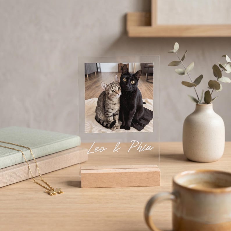 Peut inclure: Un pr&eacute;sentoir photo en acrylique transparent avec une photo de deux chats, un tabby et un noir, avec les noms "Leo & Phia" en &eacute;criture blanche. Le pr&eacute;sentoir repose sur une base en bois. Un collier dor&eacute; et des livres sont en arri&egrave;re-plan.
