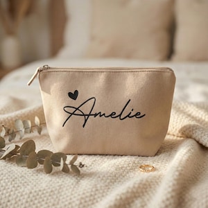 Puede incluir: Un neceser beige con cierre de cremallera. La bolsa presenta un coraz&oacute;n negro y el nombre "Amelie" en escritura cursiva. Un anillo dorado descansa sobre la superficie texturizada.