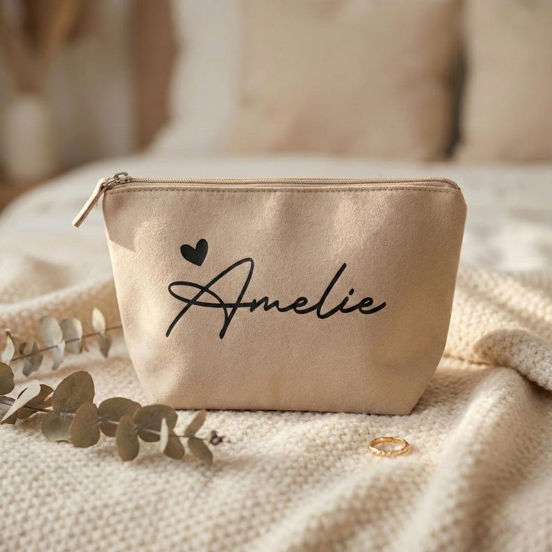 Puede incluir: Un neceser beige con cierre de cremallera. La bolsa presenta un coraz&oacute;n negro y el nombre "Amelie" en escritura cursiva. Un anillo dorado descansa sobre la superficie texturizada.