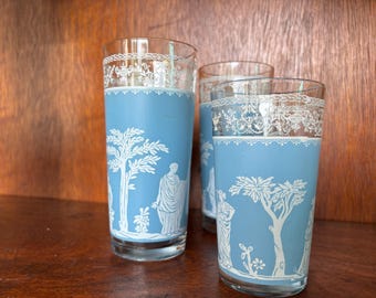 Può includere: Tre alti bicchieri trasparenti con una fascia azzurra con figure e alberi classici bianchi. I bordi hanno un design bianco e ornato. I bicchieri sono su uno sfondo di legno scuro.