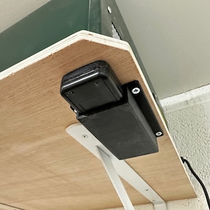 May include: A black rectangular device is mounted to a wooden surface with white metal brackets. The device is attached to a green box above the wooden surface.