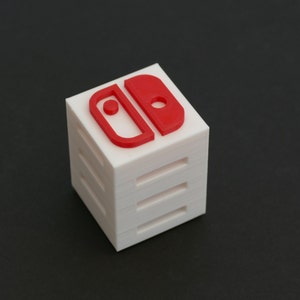 May include: A white cube with a red Nintendo Switch logo on the top. The cube is made of multiple layers.