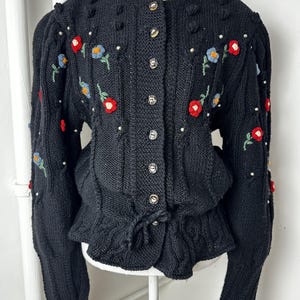 Puede incluir: Una chaqueta de punto negra con cuello redondo, mangas largas y cierre frontal de botones. La prenda está decorada con pequeñas flores bordadas en rojo y azul, tallos verdes y cuentas blancas dispersas por todo el tejido.