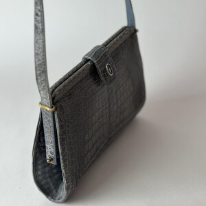 Op de afbeelding: Een grijze crossbody tas van krokodil-ge&euml;mbost leer met een klep sluiting en een zilveren knoop. De tas heeft een lange band en een korte handgreep.