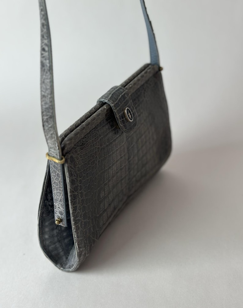 Op de afbeelding: Een grijze crossbody tas van krokodil-ge&euml;mbost leer met een klep sluiting en een zilveren knoop. De tas heeft een lange band en een korte handgreep.