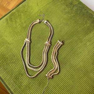 May include: A gold-toned chain necklace and bracelet set. The necklace is made of three linked chains, and the bracelet is a single chain. Both pieces have a slightly textured finish.