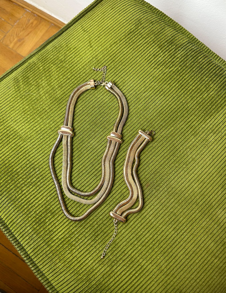 May include: A gold-toned chain necklace and bracelet set. The necklace is made of three linked chains, and the bracelet is a single chain. Both pieces have a slightly textured finish.