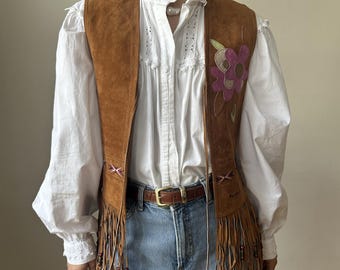 May include: A brown suede vest with fringe and a floral embroidery design. The vest has a tie closure and is worn over a white blouse and blue jeans.
