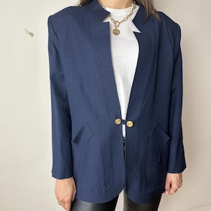 Puede incluir: Una chaqueta azul marino con cuello largo abierto y botones dorados. La chaqueta tiene un corte ajustado y está hecha de una tela texturizada.