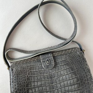 Op de afbeelding: Een grijze crossbody tas van krokodillenleer met een klep sluiting en een klein metalen logo aan de voorkant.
