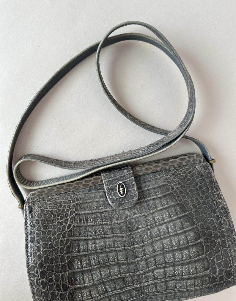 Op de afbeelding: Een grijze crossbody tas van krokodillenleer met een klep sluiting en een klein metalen logo aan de voorkant.