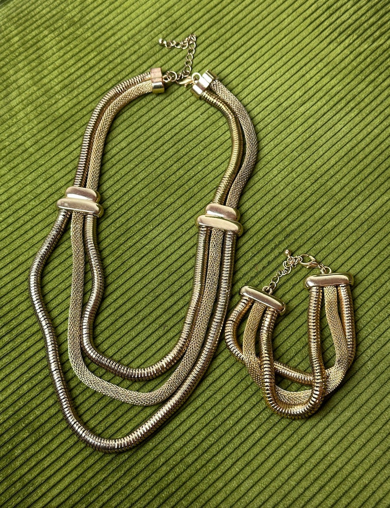 May include: A gold-tone layered chain necklace and bracelet set. The necklace has three strands of chain, and the bracelet has two strands.