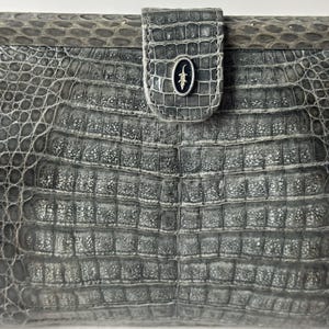 Op de afbeelding: Een grijze clutch van alligatorskin met een zilveren alligatorlogo aan de voorkant.