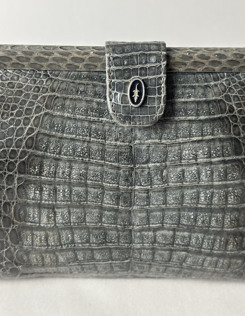 Op de afbeelding: Een grijze clutch van alligatorskin met een zilveren alligatorlogo aan de voorkant.