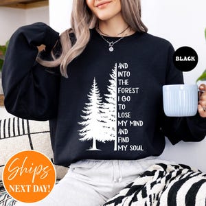 May include: A black long-sleeve sweatshirt featuring a white graphic of two pine trees and the text: AND INTO THE FOREST I GO TO LOSE MY MIND AND FIND MY SOUL. The sweatshirt has a crew neckline and is worn with light-coloured trousers.