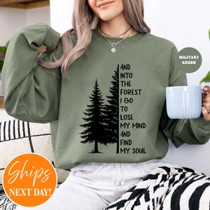 May include: A military green crewneck sweatshirt featuring a black graphic of two pine trees and the text: AND INTO THE FOREST I GO TO LOSE MY MIND AND FIND MY SOUL. The sweatshirt has long sleeves and a ribbed neckline, worn with a light-coloured garment.