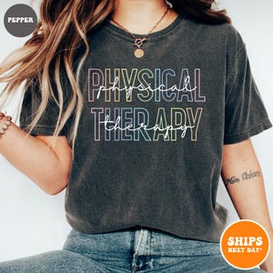 May include: A dark grey t-shirt with the text "PHYSICAL THERAPY" in rainbow colors. The text is in a script font.