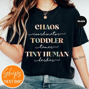 May include: A black short-sleeved t-shirt featuring the text CHAOS coordinator TODDLER tamer TINY HUMAN herder in alternating light green and peach colors. The shirt has a crew neckline and is worn with blue denim jeans and a gold necklace.