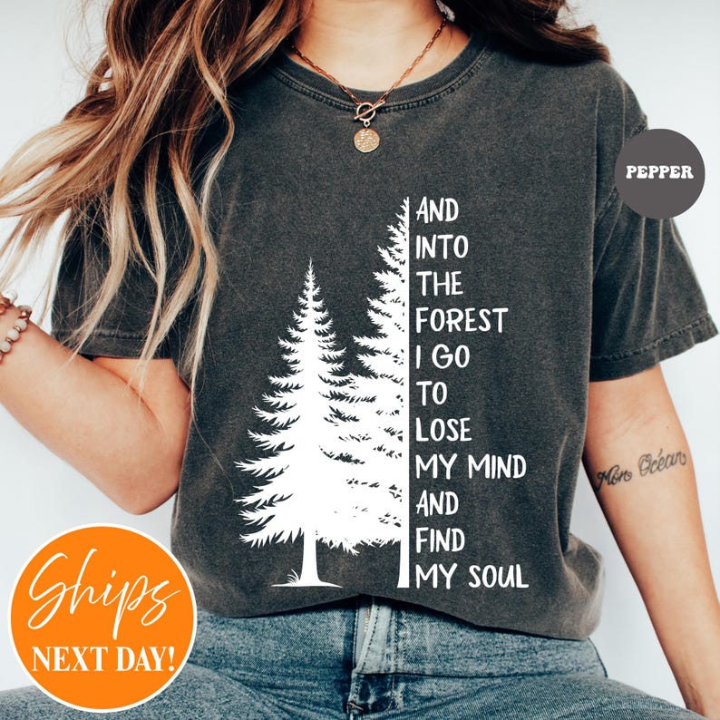 May include: A charcoal gray short-sleeve t-shirt featuring a white graphic of two pine trees and the text: AND INTO THE FOREST I GO TO LOSE MY MIND AND FIND MY SOUL. The shirt has a crew neckline and a vintage-style washed fabric appearance.