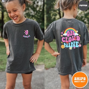May include: Dark gray t-shirt with a colorful design. The front features the number 9 with the name "Ashley" in a playful font. The back displays a rainbow, the words "On Cloud Nine," and smiley faces.