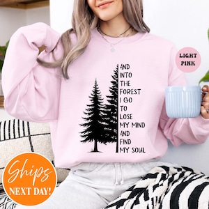 May include: A light pink crewneck sweatshirt featuring a black graphic of two pine trees and the text: AND INTO THE FOREST I GO TO LOSE MY MIND AND FIND MY SOUL. The long-sleeved garment has a ribbed collar, cuffs, and hem, with a soft, solid-coloured fabric.