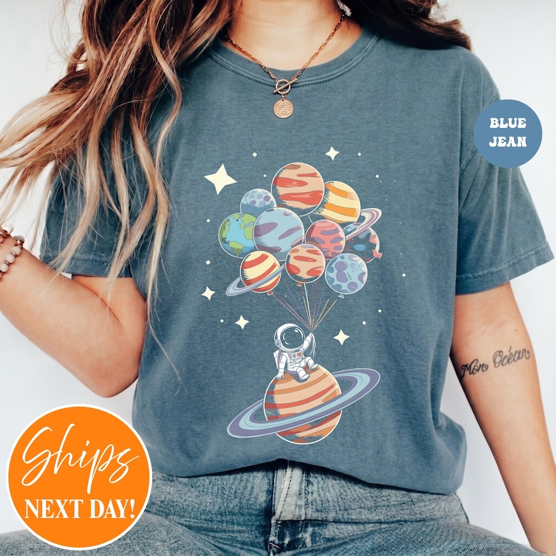 May include: A slate blue t-shirt featuring a graphic of an astronaut sitting on a ringed planet, holding a cluster of colorful planet-shaped balloons. The design includes small white stars scattered around the central space-themed illustration.