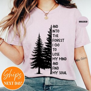 May include: A light orchid-colored t-shirt featuring a black graphic of two pine trees and the text: AND INTO THE FOREST I GO TO LOSE MY MIND AND FIND MY SOUL. The shirt has a crew neckline and short sleeves, with a small circular label reading ORCHID.