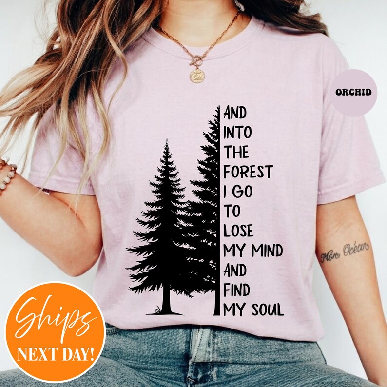 May include: A light orchid-colored t-shirt featuring a black graphic of two pine trees and the text: AND INTO THE FOREST I GO TO LOSE MY MIND AND FIND MY SOUL. The shirt has a crew neckline and short sleeves, with a small circular label reading ORCHID.