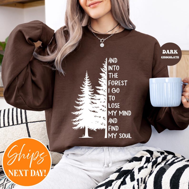May include: A dark chocolate brown crewneck sweatshirt featuring a white graphic of two pine trees and the text: AND INTO THE FOREST I GO TO LOSE MY MIND AND FIND MY SOUL. The sweatshirt has long sleeves and a ribbed collar, cuffs, and hem.